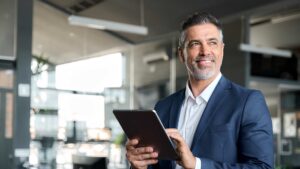 A man in a blue suit holds a clipboard in an office and smiles