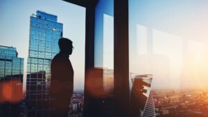 A man wearing glasses and a suit looks of out an office window at the sunset over a city
